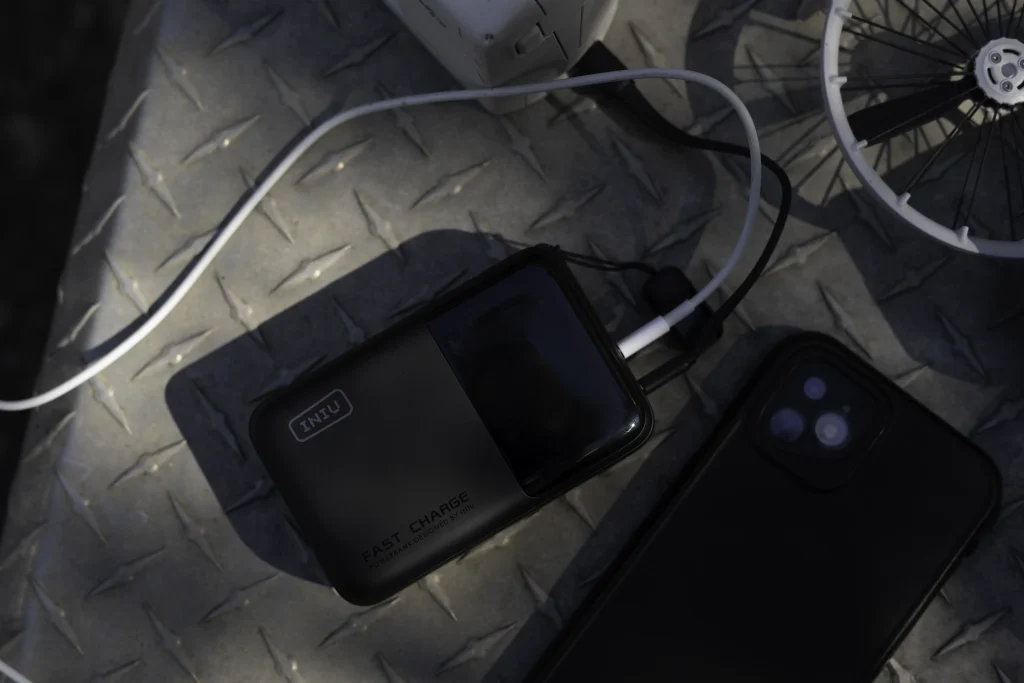 Image of the INIU P63 100W 25 000 mAh charger with a drone, drone batteries, and a black iPhone. These are all on an aluminum checkerplate surface.