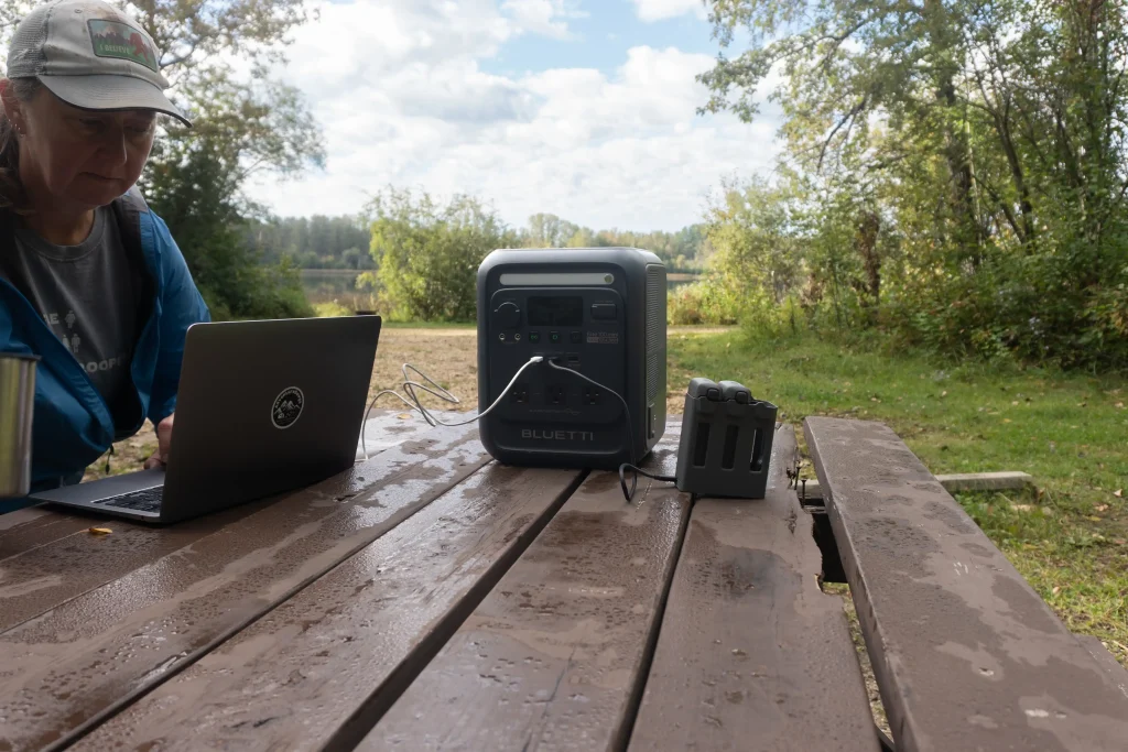 A BLUETTI Elite 100 Mini power station is on a picnic table in a campsite. There is a woman sitting on the left working on a laptop that is plugged in and a set of batteries is plugged in on the right side.
