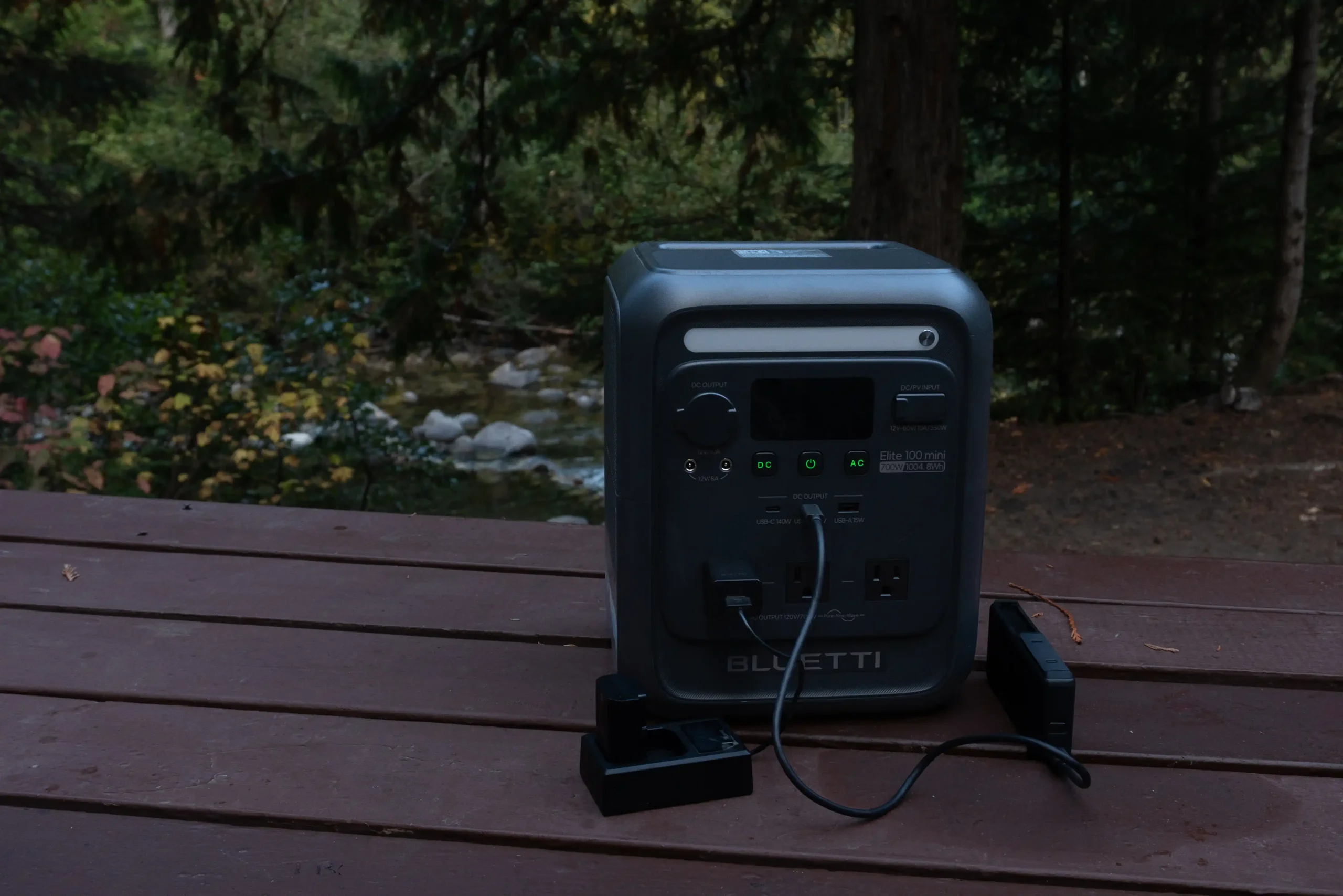 A BLUETTI Elite 100 mini is on a wooden table in a wooded area. There are batteries plugged in to charge.